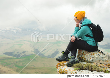 Young woman with a backpack pensively sitting on the edge of a rock and looking at the sky with 42578782