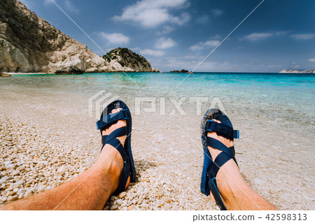 Young male feets wear blue flip-flop sandal sunbathing on sea beach with clear water 42598313