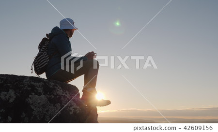 A traveler drinks tea from a mug at the peak of the mountain. Sits on a rock, in the background a 42609156