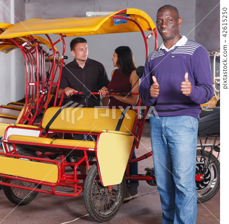 Smiling African-American pedicab driver standing near rickshaw cycle Smiling African-American pedicab driver standing near rickshaw cycle 42615250