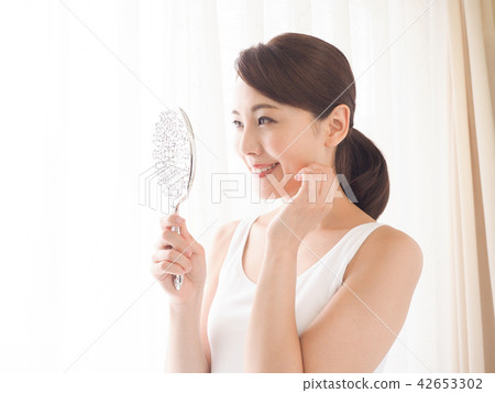 A woman looking at a mirror at the window side with his hand on his cheek 42653302