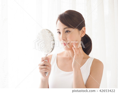 A woman looking at a mirror at the window side with his hand on his cheek 42653422
