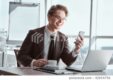Young businessman in eyeglasses at office sitting drinking coffe Young businessman in eyeglasses at office sitting drinking coffe 42654756