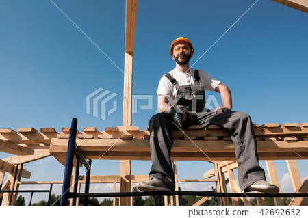 The man builder sits on the edge of the roof of the frame house, in a yellow helmet and gray 42692632