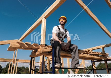 The man builder sits on the edge of the roof of the frame house, in a yellow helmet and gray 42692666