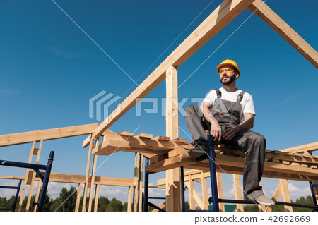 The man builder sits on the edge of the roof of the frame house, in a yellow helmet and gray 42692669