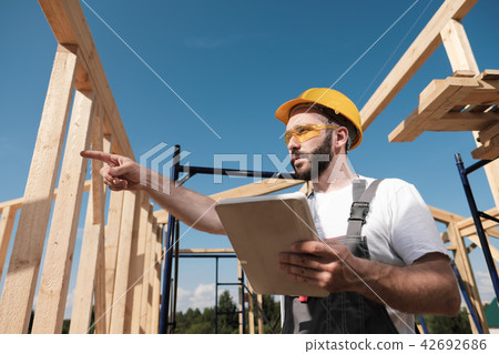 The man is a builder on the background of the roof of a frame house, in a yellow helmet and gray The man is a builder on the background of the roof of a frame house, in a yellow helmet and gray 42692686