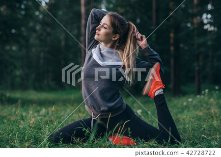 Young woman practicing yoga doing one-legged pigeon pose sitting on grass in park 42728722