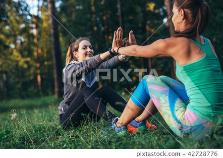 Two positive girlfriends doing partner sit-up exercise with high-five in summer park Two positive girlfriends doing partner sit-up exercise with high-five in summer park 42728776