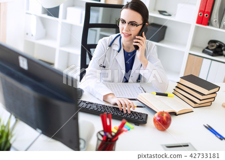A beautiful young girl in a white robe is sitting at the table, talking on the phone and holding her 42733181