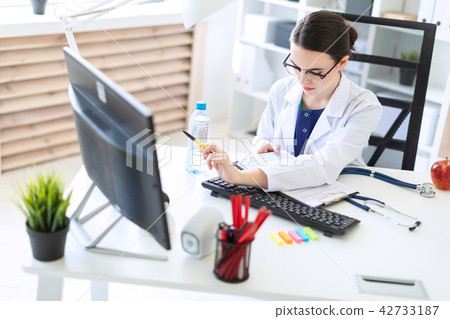 A beautiful young girl in a white robe is sitting at a computer desk with documents and a pen in her 42733187