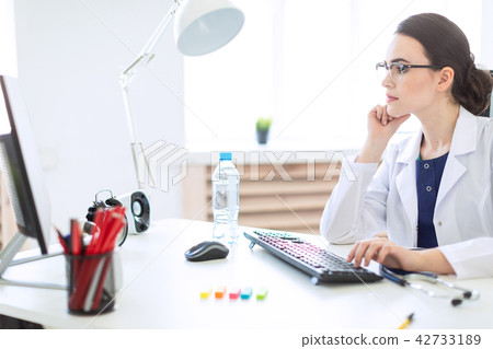 A beautiful young girl in a white robe is sitting at the table and typing on the keyboard. 42733189