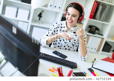 Beautiful young girl sitting in headphones at desk in office, eating yogurt and looking at monitor. 42733194