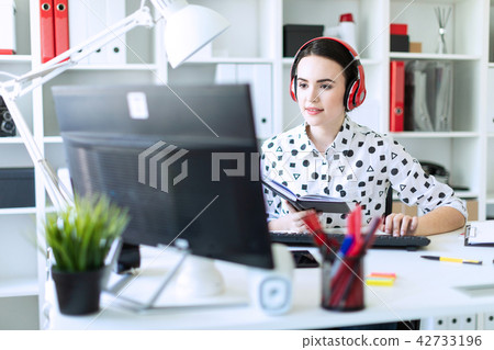 Beautiful young girl sits in headphones at desk in office, holds a notebook in her hand and prints 42733196