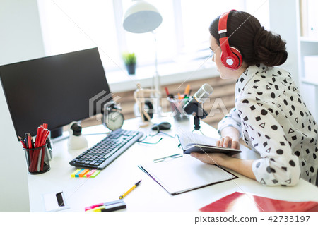 Beautiful young girl sits in headphones and with a notepad at the table in the office and speaks 42733197