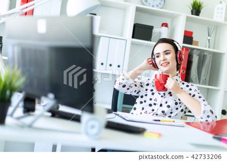 A young girl sits in headphones at a table in the office, holds a red cup in her hands and looks at 42733206