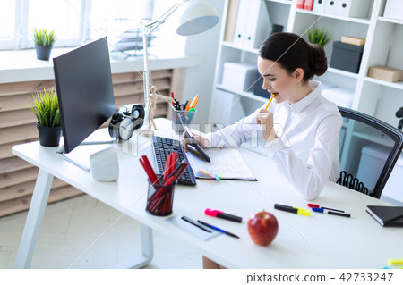 A young girl in the office holds a pen in her mouth and works with a calculator, documents and a A young girl in the office holds a pen in her mouth and works with a calculator, documents and a 42733247