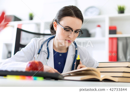 A beautiful young girl in a white robe is sitting at the computer desk with a marker and a book. A 42733844