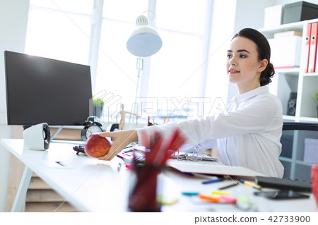 A young girl at the office is working with documents and stretches forth an apple. 42733900
