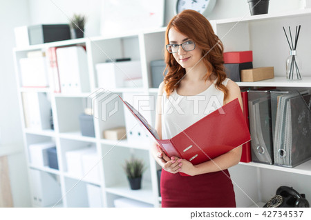A beautiful young girl stands near a stack in the office and holds a folder with documents in her 42734537
