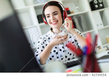 Beautiful young girl sitting in headphones at desk in office, eating yogurt and looking at monitor. 42734554