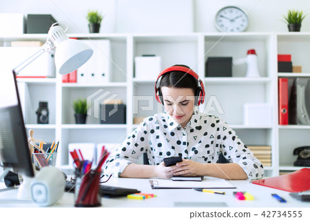 Beautiful young girl sitting in headphones at desk in office and holding a phone. 42734555
