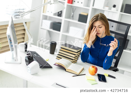 A young girl is sitting at a computer desk, holding glasses in her hand and rubbing her eyes. Before 42734716