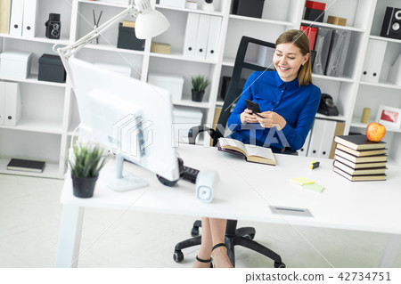 A young girl sits at a computer desk and holds a phone in her hand. Before the girl lies an open A young girl sits at a computer desk and holds a phone in her hand. Before the girl lies an open 42734751