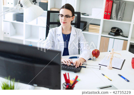 A beautiful young girl in a white robe is sitting at the table and typing on the keyboard. 42735229