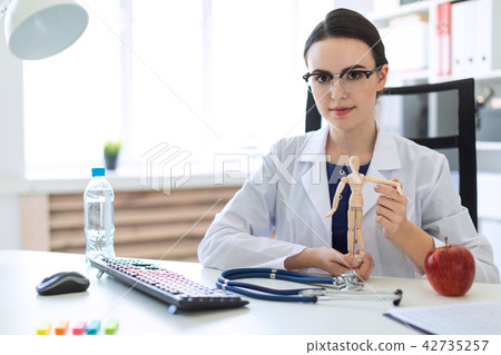 A beautiful young girl in a white robe is sitting at a table and holding a wooden figure of a man in 42735257