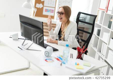 A young girl is sitting at the table in the office, looking at the monitor and waving her hand. 42735299