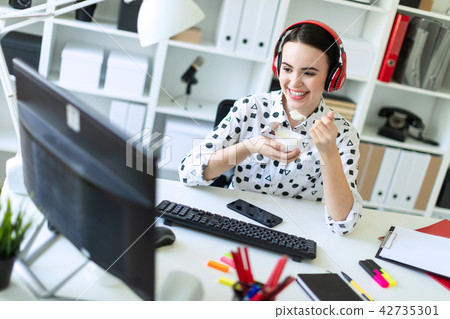 Beautiful young girl sitting in headphones at desk in office, eating yogurt and looking at monitor. 42735301