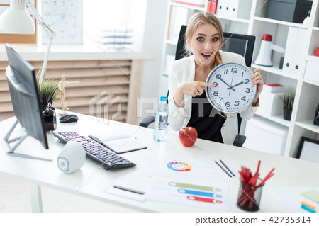 A young girl sits at a table in the office and points a finger at the clock face. Before the girl on 42735314