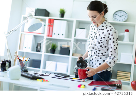 A young girl stands in the office near the table and pours coffee from the coffee pot into a red cup 42735315