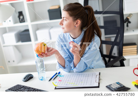 A young girl sits at a table in the office and holds an apple in her hand. A young girl sits at a table in the office and holds an apple in her hand. 42755780