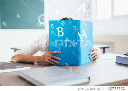 Student girl covering her face behind a book in classroom. 42777532