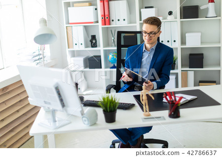 A young man sits at a table in the office and holds documents and a pen in his hands. 42786637