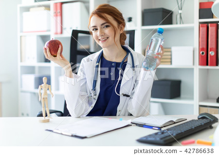 A beautiful young girl in a white robe sits at a table in the office and holds an apple and a bottle 42786638