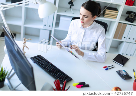 A young girl in the office holds a pen in her hand and works with documents and a computer. 42786747
