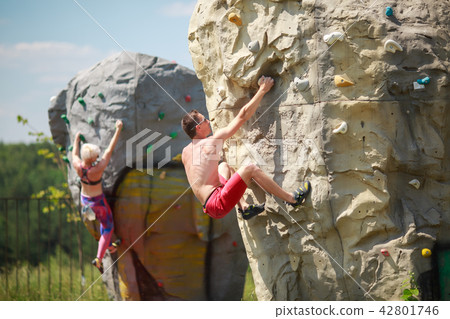 Photo of sports man in red shorts and woman on workout at climbing boulder against blue sky with 42801746