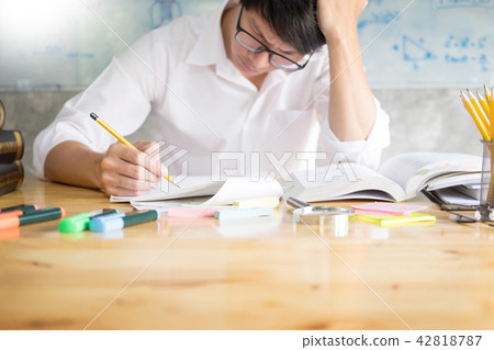 teenage man sitting by wooden table reading and writting on note teenage man sitting by wooden table reading and writting on note 42818787