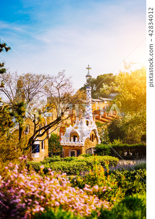 Colorful mosaic building in Park Guell in warm sunset light. Framed by garden flowers in foreground Colorful mosaic building in Park Guell in warm sunset light. Framed by garden flowers in foreground 42834012