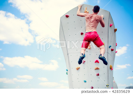 Photo from back of athlete man in red shorts practicing on wall for climbing against blue sky with Photo from back of athlete man in red shorts practicing on wall for climbing against blue sky with 42835629