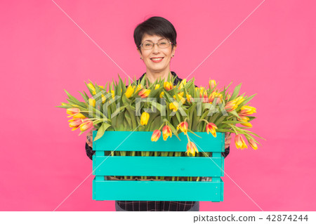 Floristics, holidays and people concept - Middle-aged woman holding box of tulips on a pink 42874244