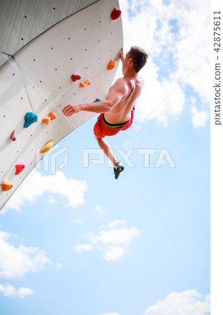 Photo from bottom of young athletic man exercising on wall for climbing against blue sky with clouds 42875611