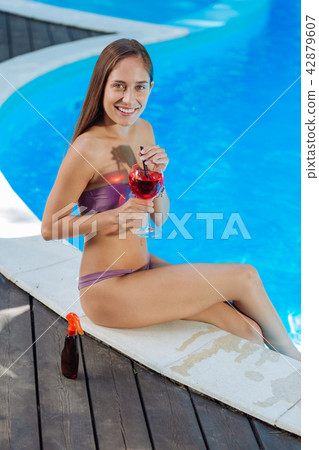 Young woman sitting near outside pool and drinking cold drink Young woman sitting near outside pool and drinking cold drink 42879607