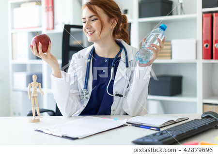 A beautiful young girl in a white robe sits at a table in the office and holds an apple and a bottle A beautiful young girl in a white robe sits at a table in the office and holds an apple and a bottle 42879696