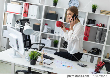 Young girl sitting at desk in office, holding a red cup in her hand and talking on the phone. 42879843