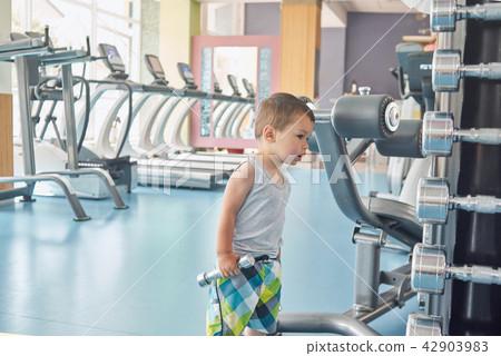 Little child standing in gym in front of metallic dumbbells and racetracks. Little child standing in gym in front of metallic dumbbells and racetracks. 42903983