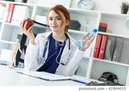 A beautiful young girl in a white robe sits at a table in the office and holds an apple and a bottle 42904055